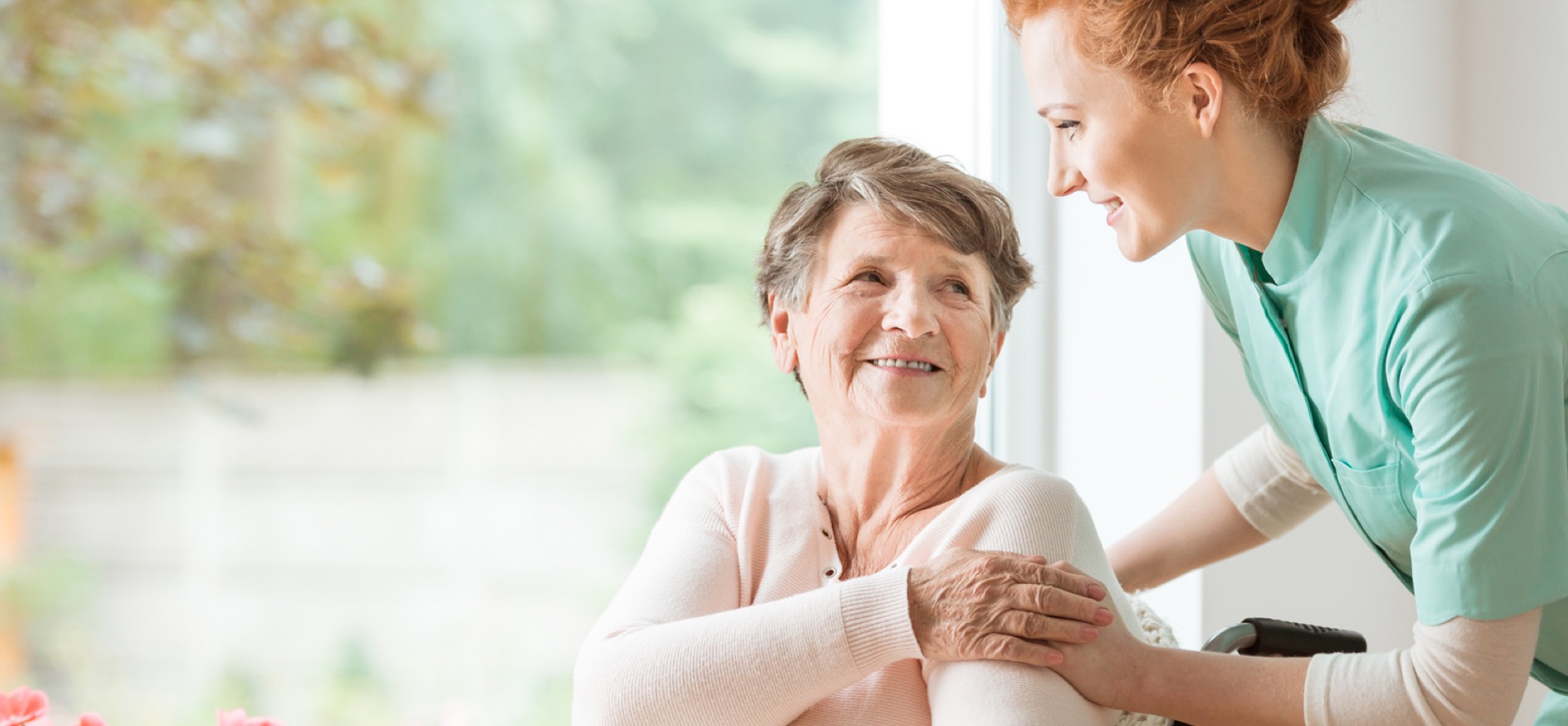 Betreuerin und Seniorin im vertrauten Gespräch am Fenster