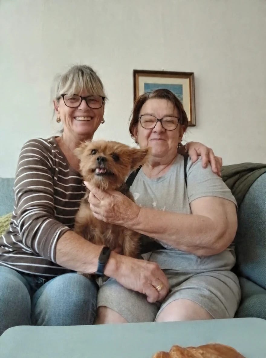 Eine Betreuerin und eine Kundin sitzen nebeneinander auf dem Sofa, zwischen ihnen ein kleiner Hund