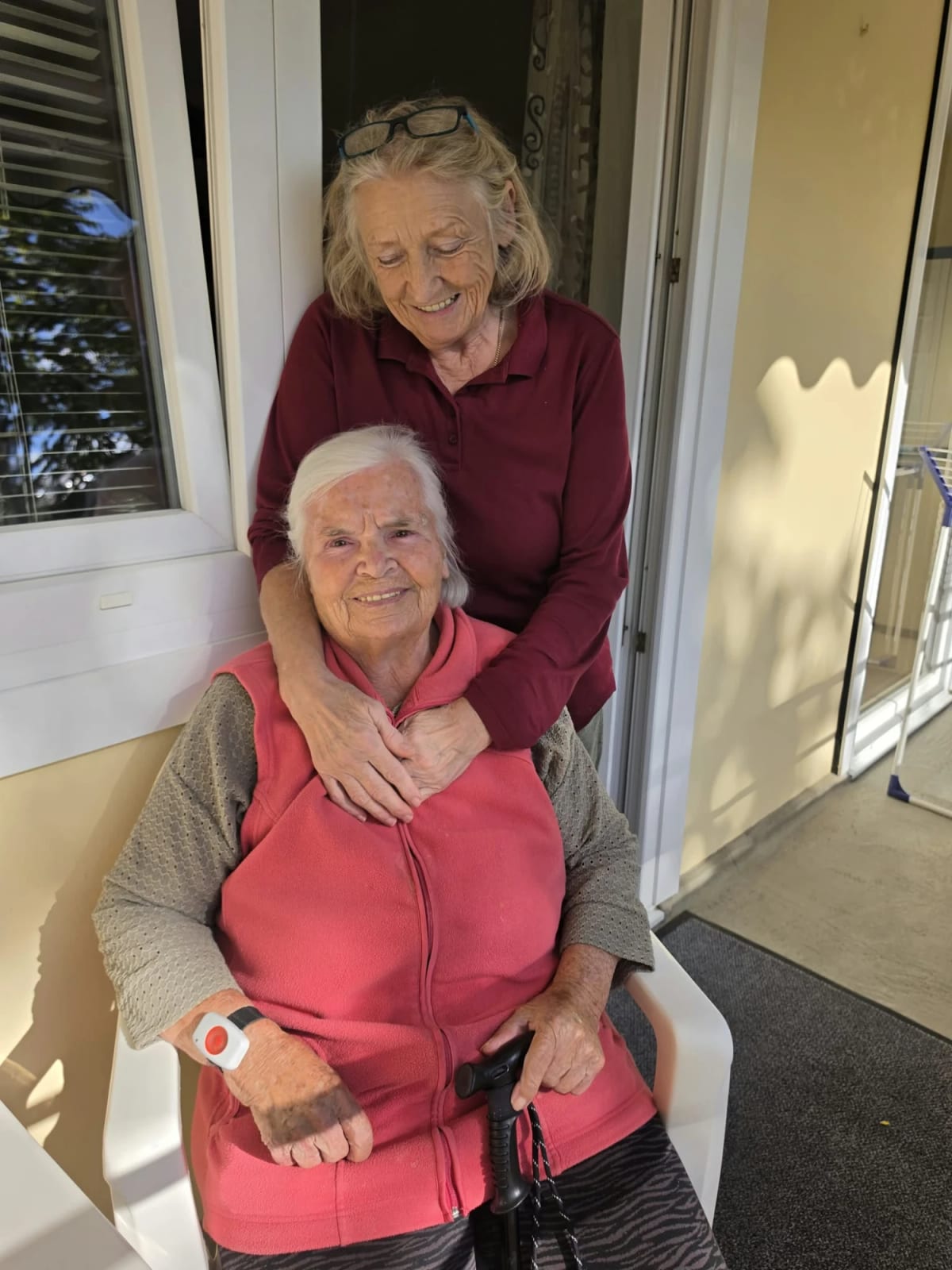 Eine Betreuerin steht hinter einer sitzenden Seniorin auf dem Balkon und legt ihr die Arme um die Schultern; die Seniorin trägt einen Hausnotruf am Handgelenk