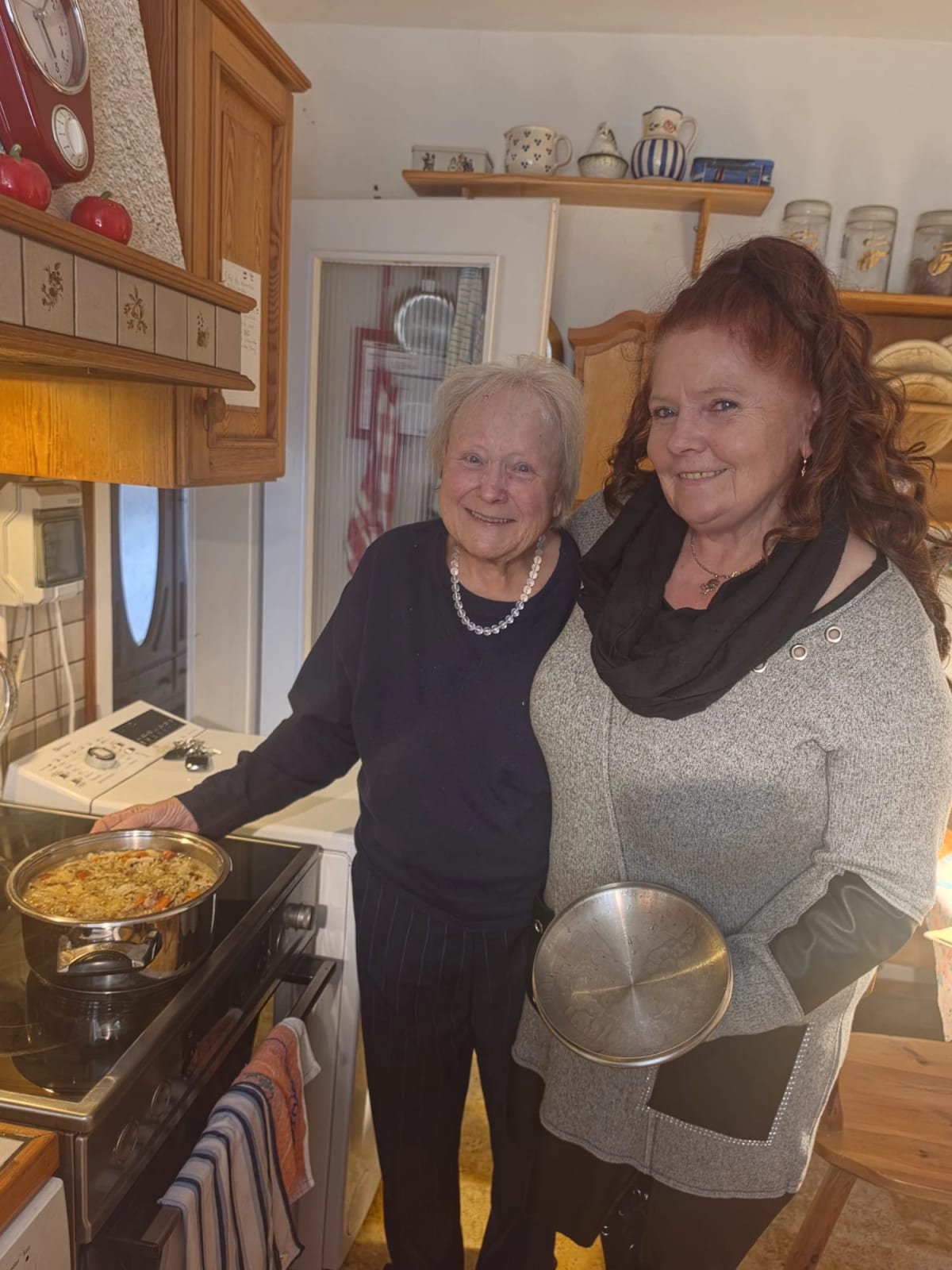 Eine Seniorin und ihre Betreuerin stehen lachend in der Küche am Herd, auf der Platte steht ein Topf mit fertigem Essen