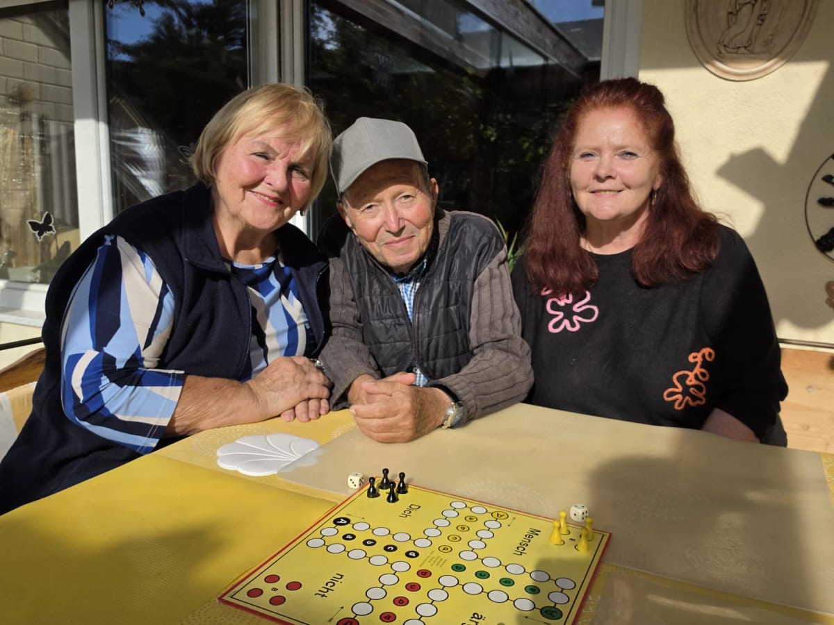 Zwei Betreuerinnen und ein Senior sitzen in einem sonnigen Wintergarten am Tisch und spielen ein Brettspiel
